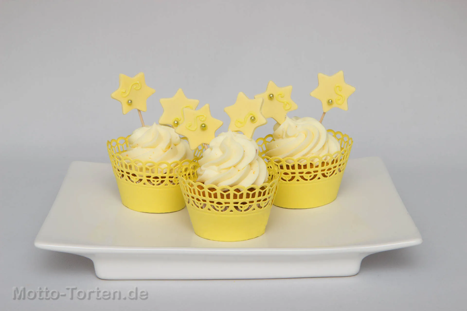 Drei Cupcakes mit hellem Frischkäse-Topping in gelben Manschetten, dekoriert mit Sternen aus Fondant auf Stäbchen