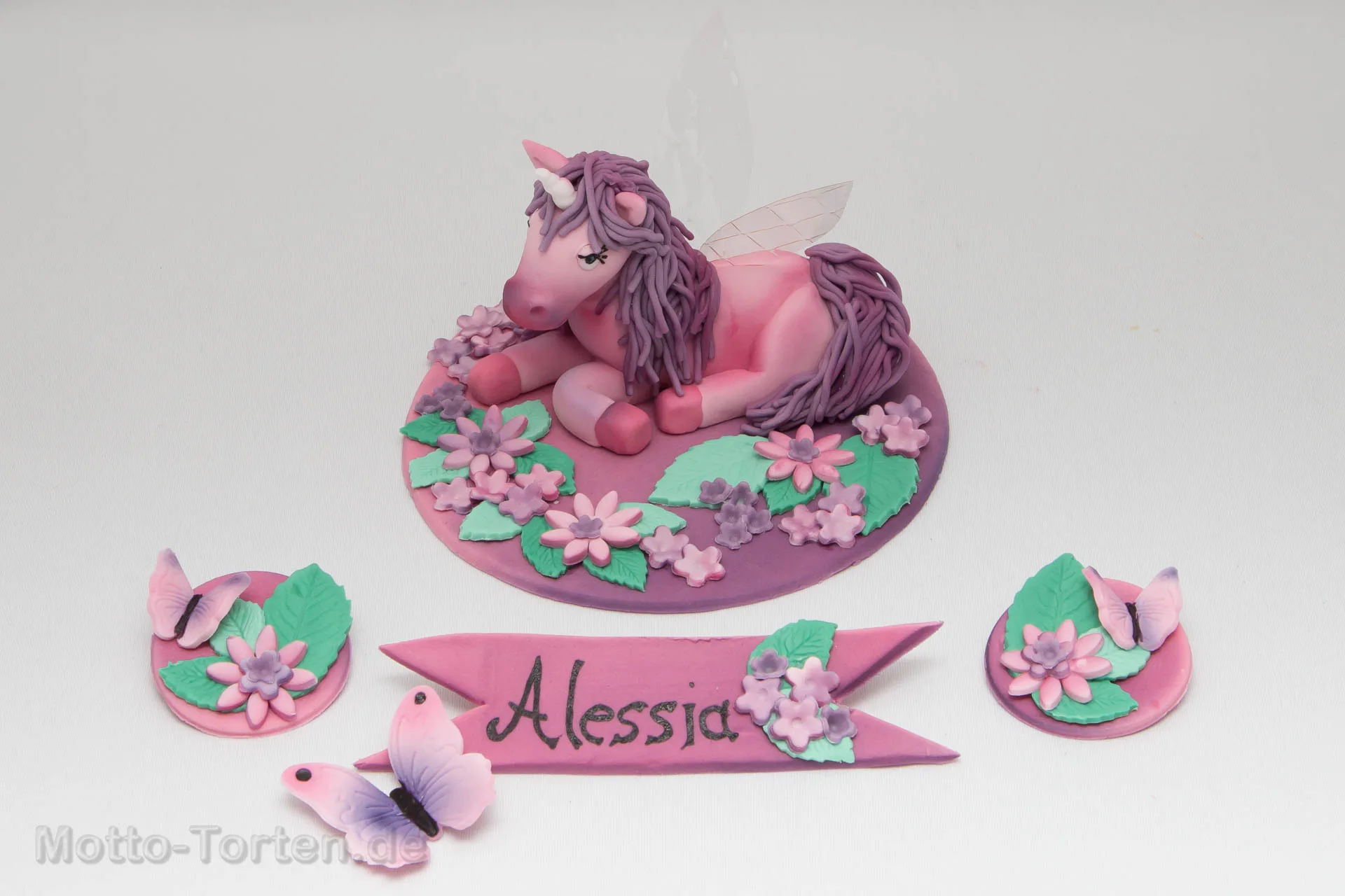 Handmodelliertes rosa Einhorn aus Fondant mit Flügeln aus Gelatine, auf einer Bodenplatte mit Blumen und Namensschild