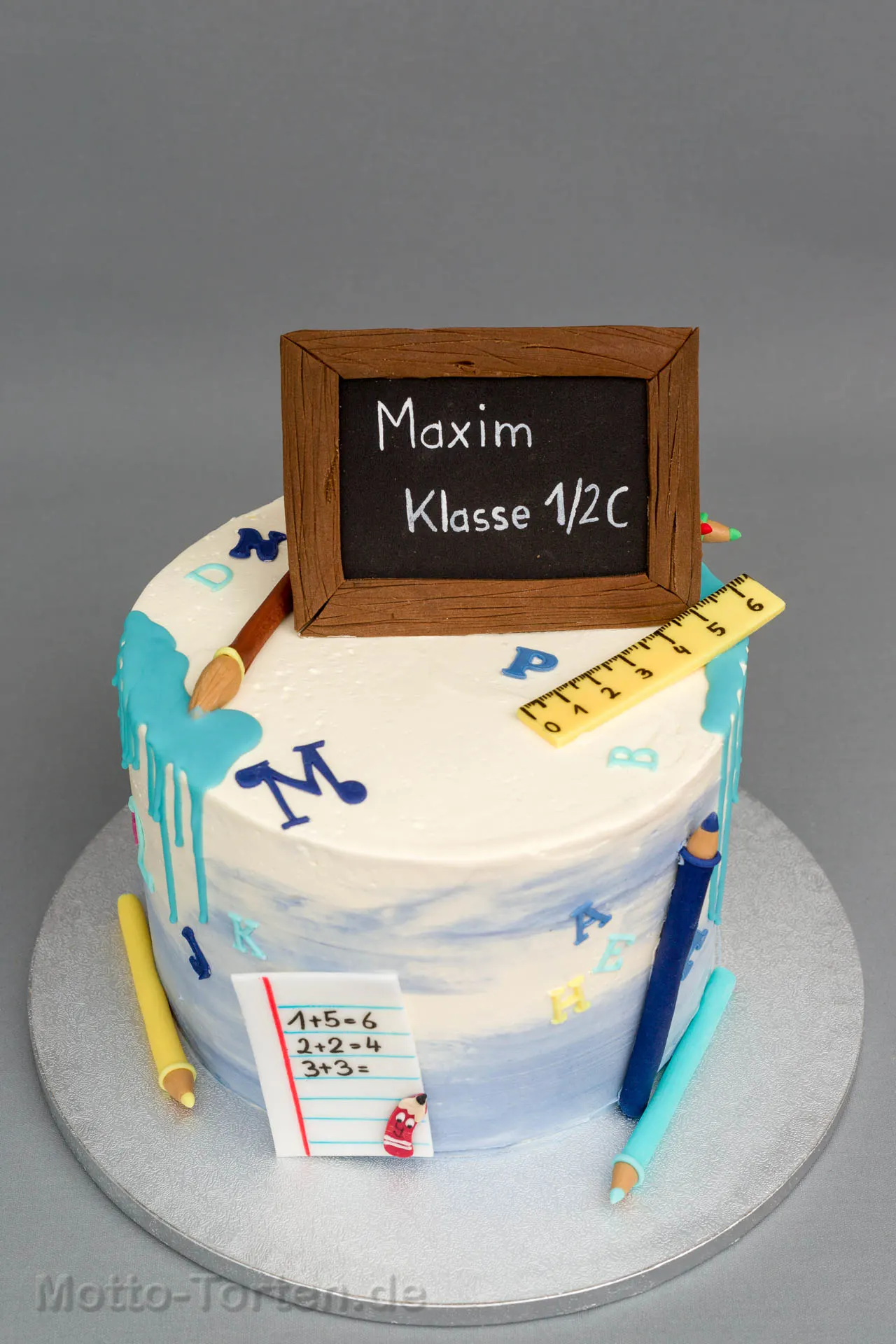 Runde Buttercremetorte in Weiß und Blau mit türkisem Drip, Fondantstiften, Lineal und einer Tafel mit Namen und Klasse