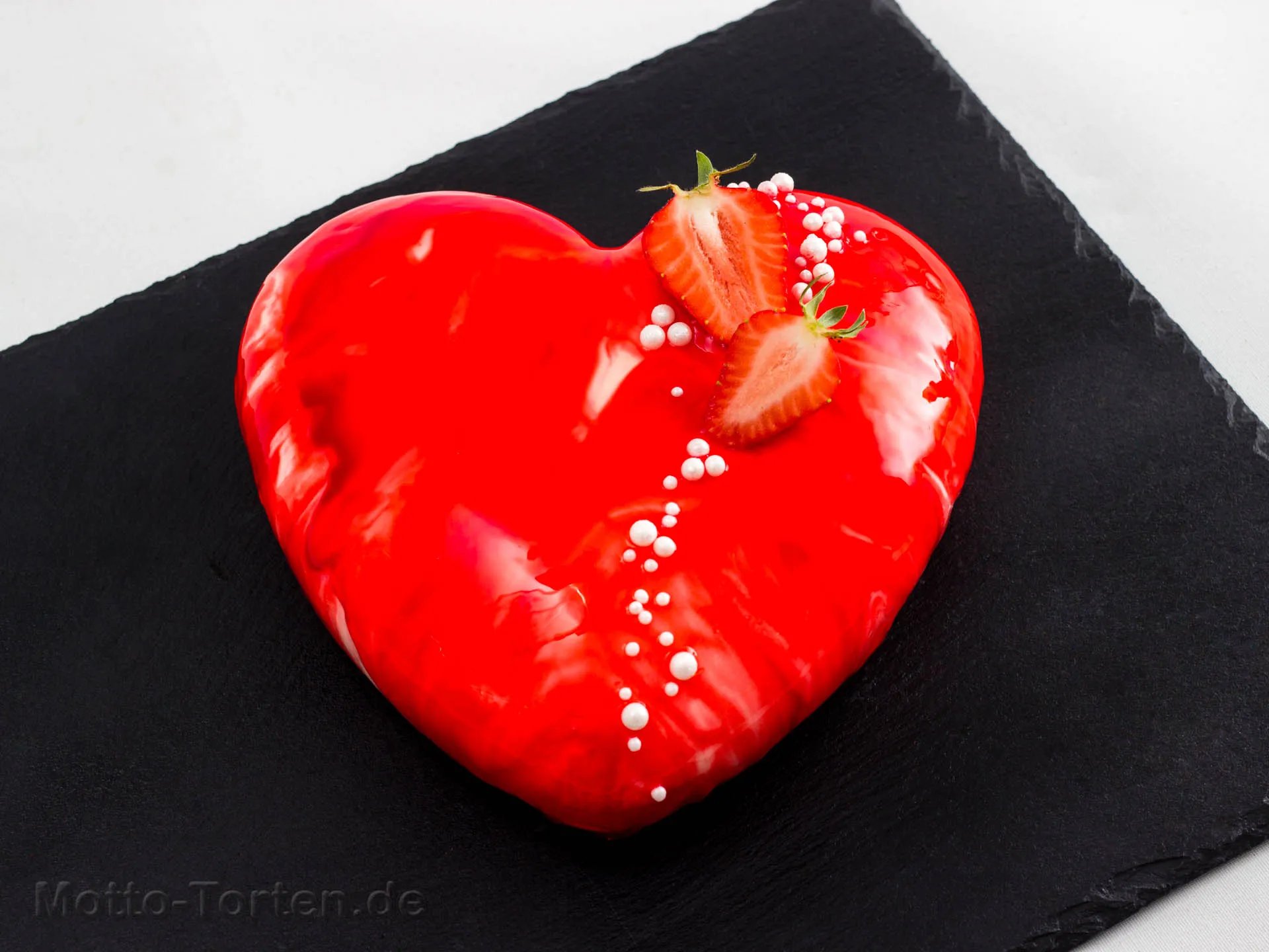 Herzförmige Torte mit spiegelnder roter Glasur auf einer Schieferplatte, garniert mit halbierten Erdbeeren und weißen Zuckerperlen