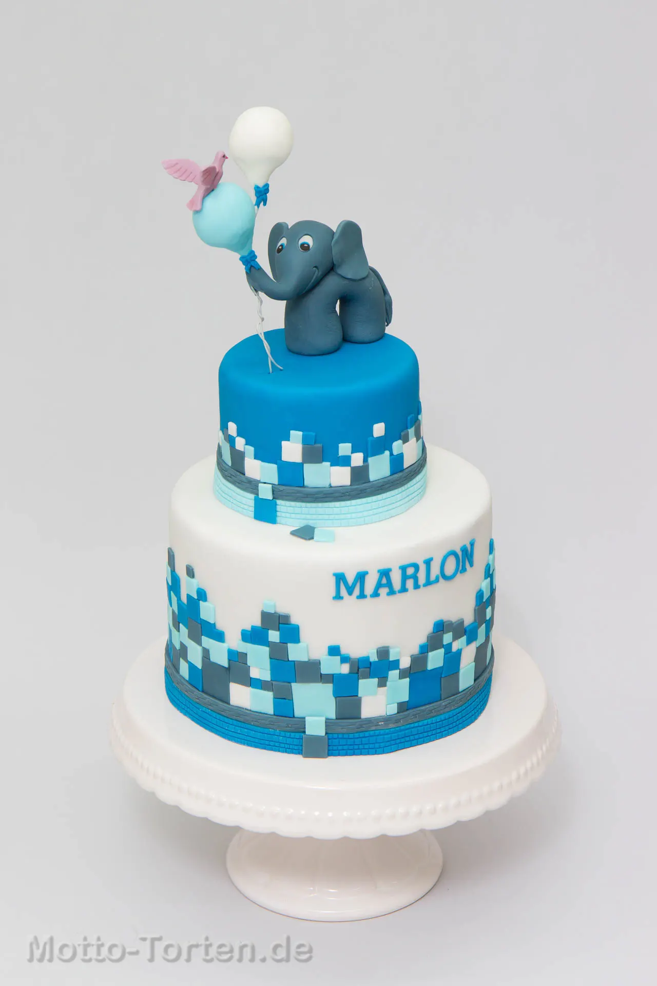 Zweistöckige blaue Babytorte mit aufgesetzten Quadraten, Namensschriftzug und einem Fondant-Elefanten mit Luftballons obenauf
