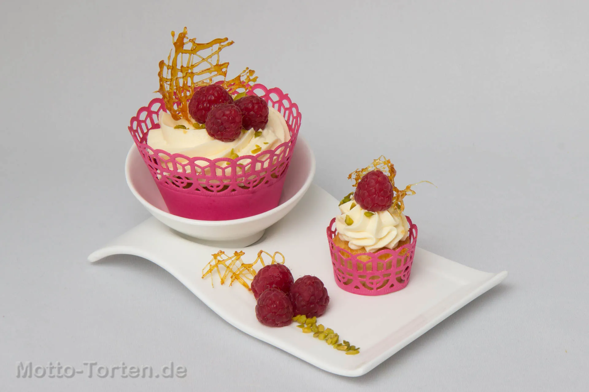 Zwei Cupcakes mit hellem Mascarpone-Topping in pinken Manschetten, garniert mit frischen Himbeeren, Pistazien und Karamellgespinst