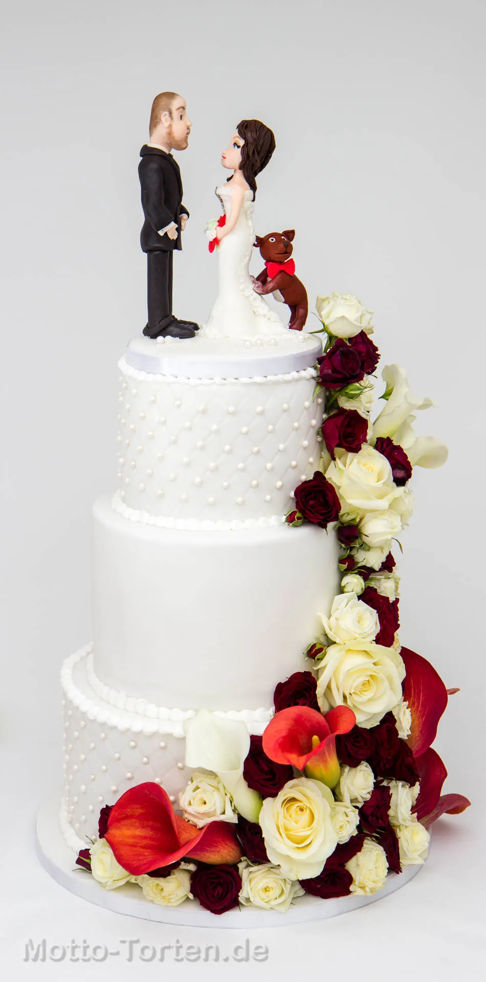 Dreistöckige weiße Hochzeitstorte mit Steppmuster und Zuckerperlen, einer Kaskade aus weißen und dunkelroten Rosen und Callas, oben das Brautpaar mit Hund aus Fondant