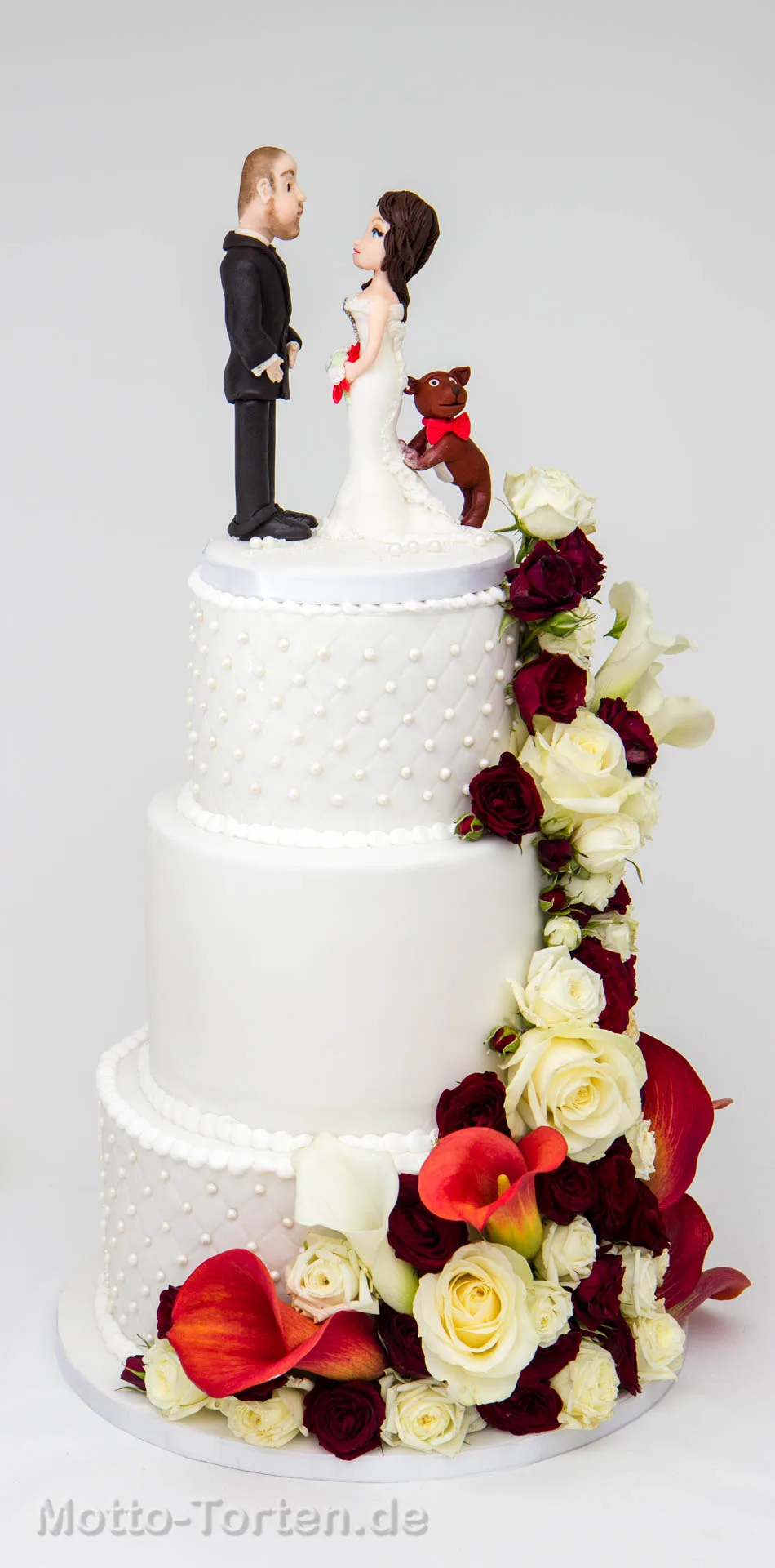 Dreistöckige weiße Hochzeitstorte mit Steppmuster und Zuckerperlen, einer Kaskade aus weißen und dunkelroten Rosen und Callas, oben das Brautpaar mit Hund aus Fondant
