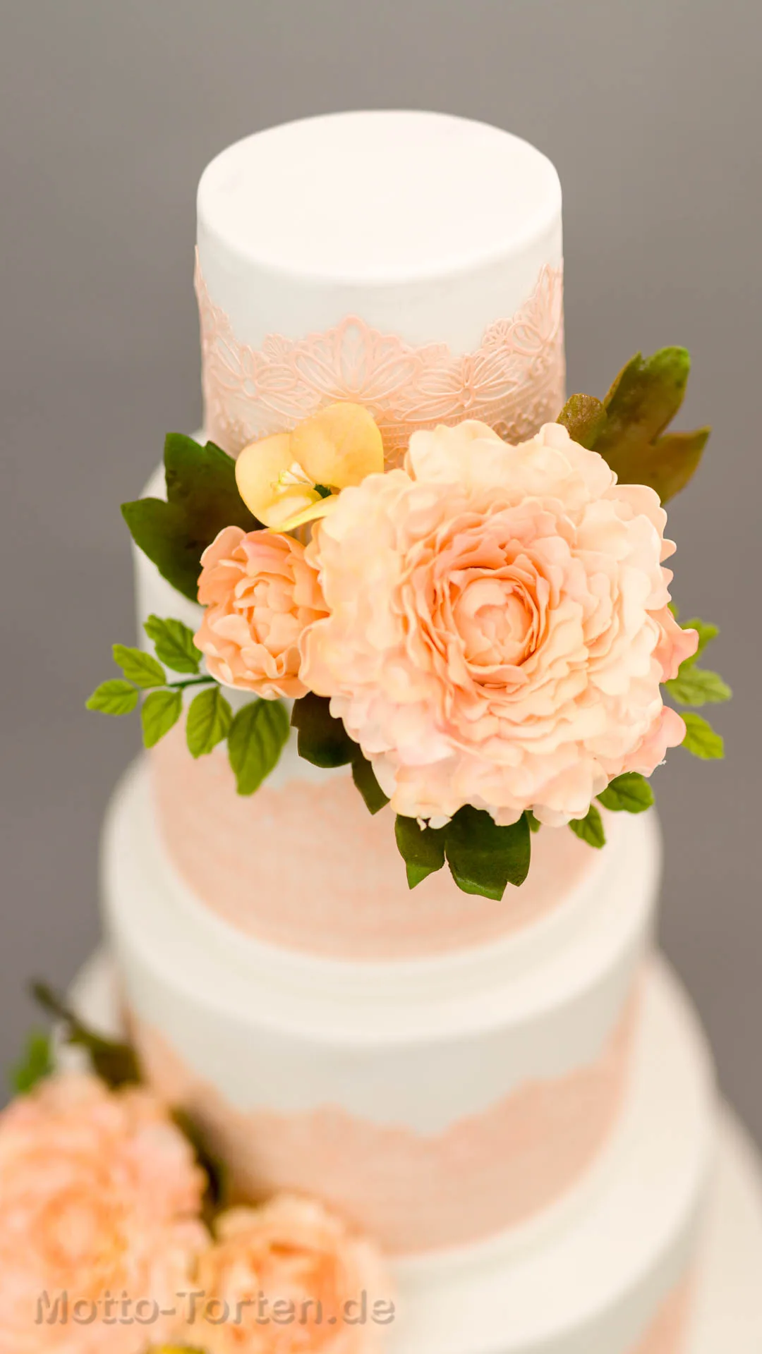 Mehrstöckige weiße Hochzeitstorte mit apricotfarbener essbarer Spitze und handmodellierten Pfingstrosen in Apricot mit grünen Blättern