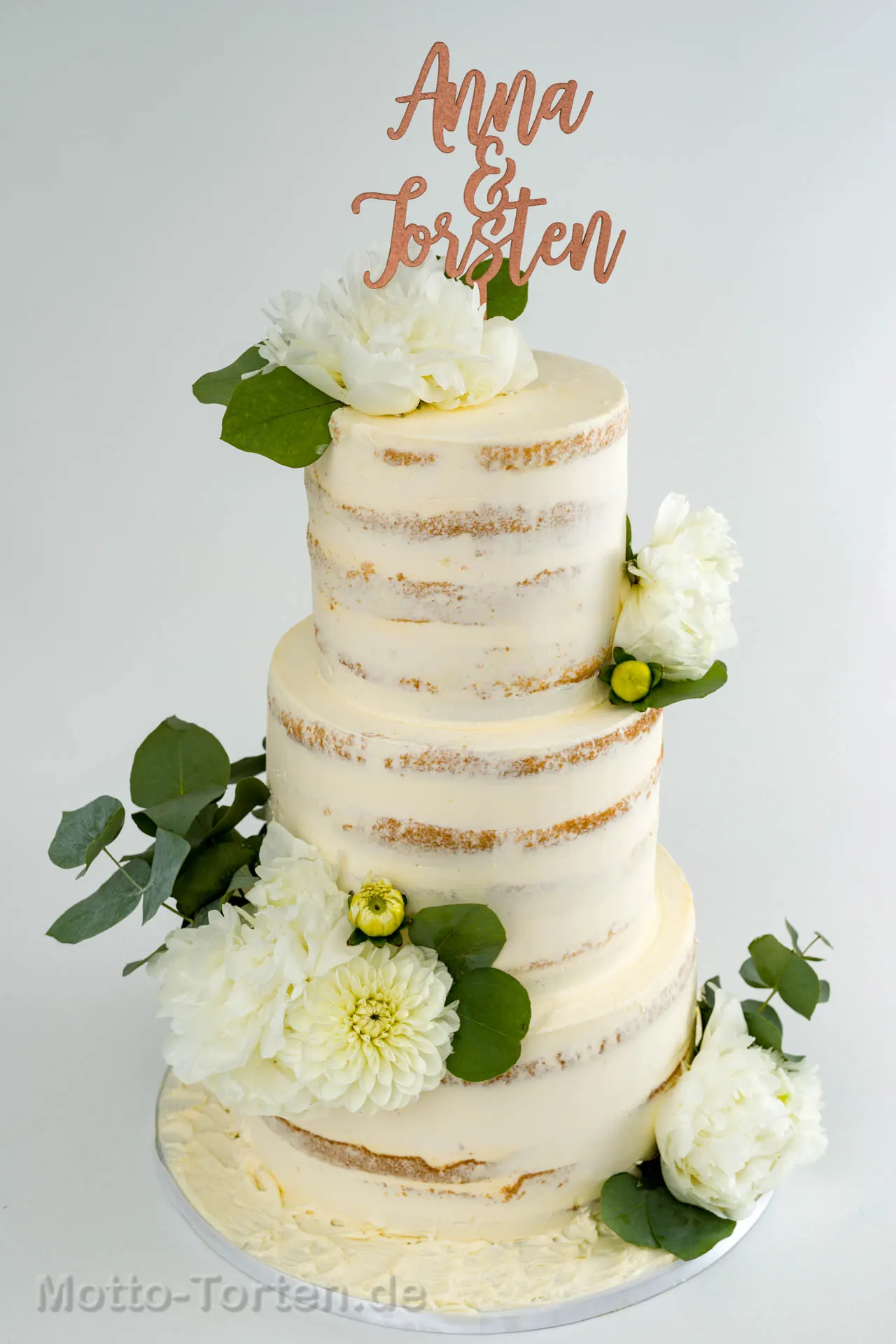 Dreistöckige Half-Naked-Hochzeitstorte mit weißen Dahlien, Pfingstrosen und Eukalyptus, obenauf ein roségoldener Caketopper