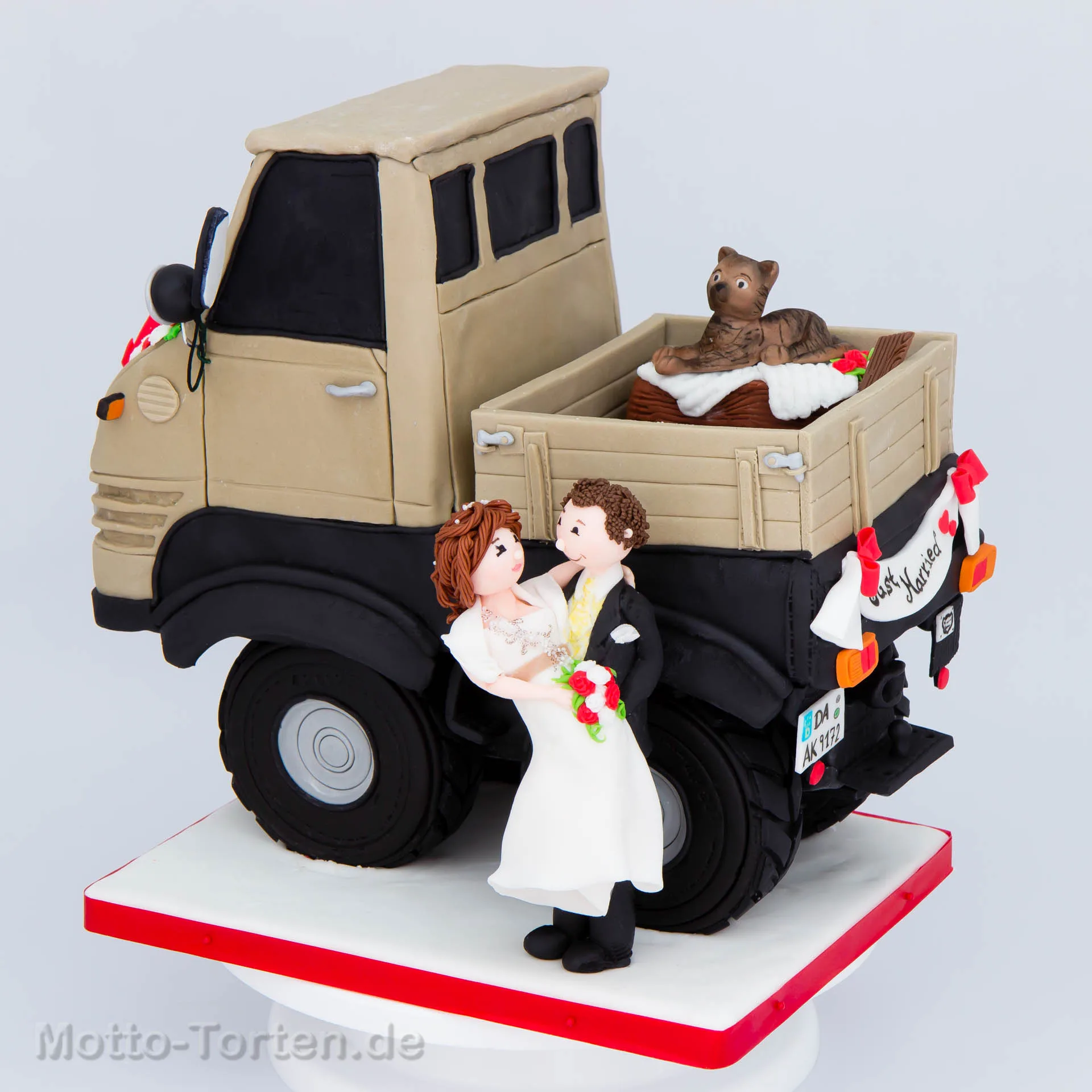 Caketopper aus Fondant: beiger Unimog mit Just-Married-Schild, davor das Brautpaar im Comicstil, auf der Ladefläche die Katze in einem Korb