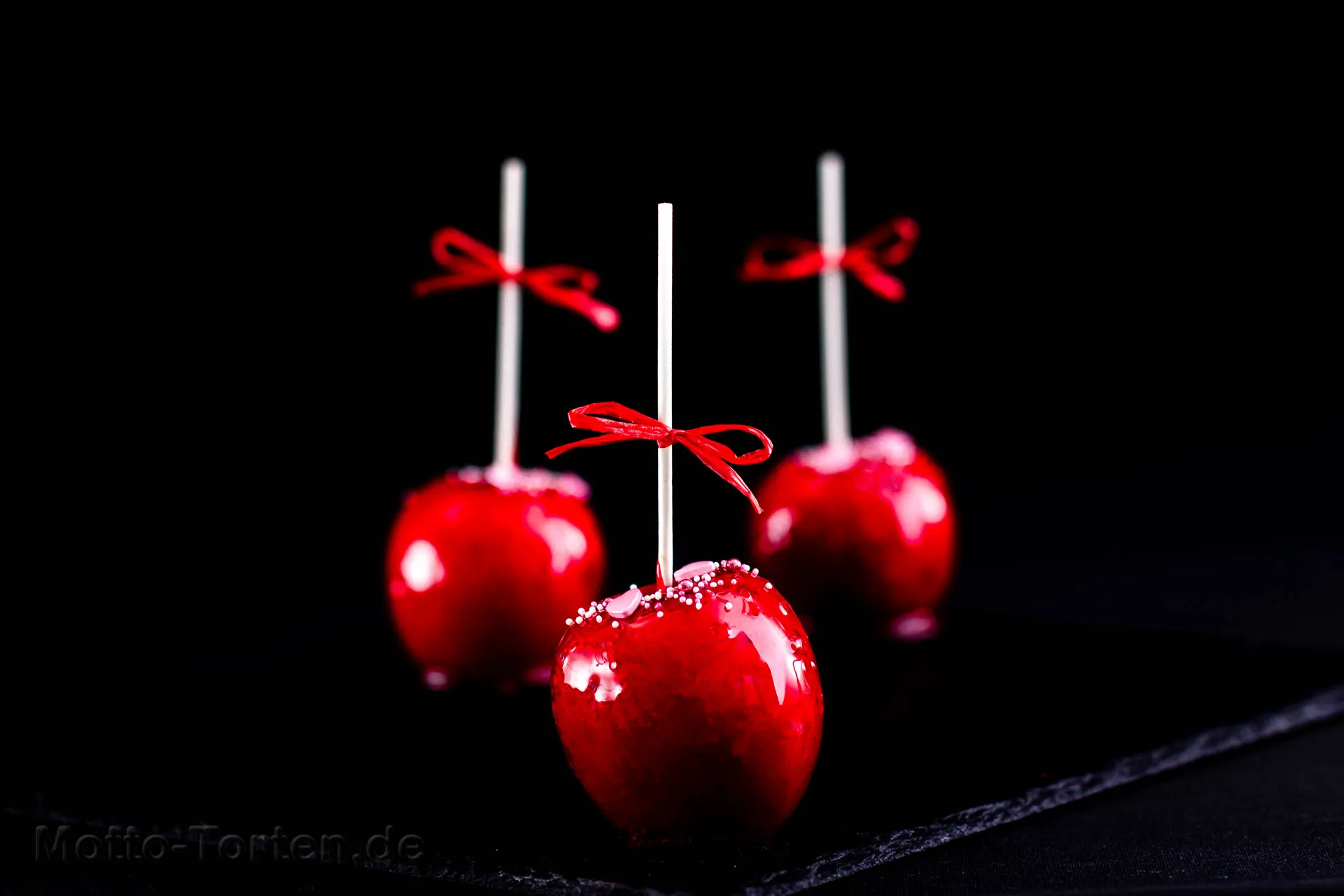 Drei rote Liebesäpfel an weißen Stielen mit roten Schleifen, glänzend kandiert und mit Zuckerperlen bestreut, vor dunklem Hintergrund