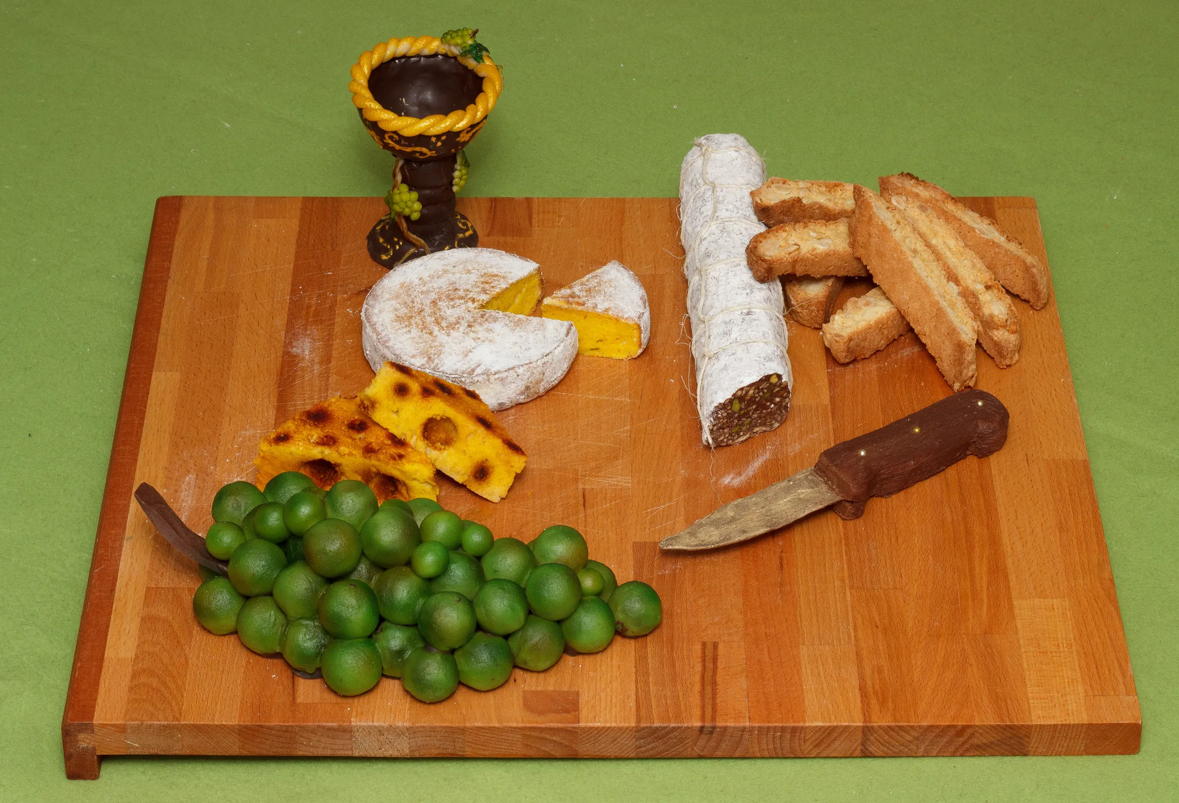 Holzbrett mit einer Vesperplatte, bei der alles aus Süßem besteht: Käselaib, Salami, Weintrauben, Cantuccini und ein Messer aus Schokolade