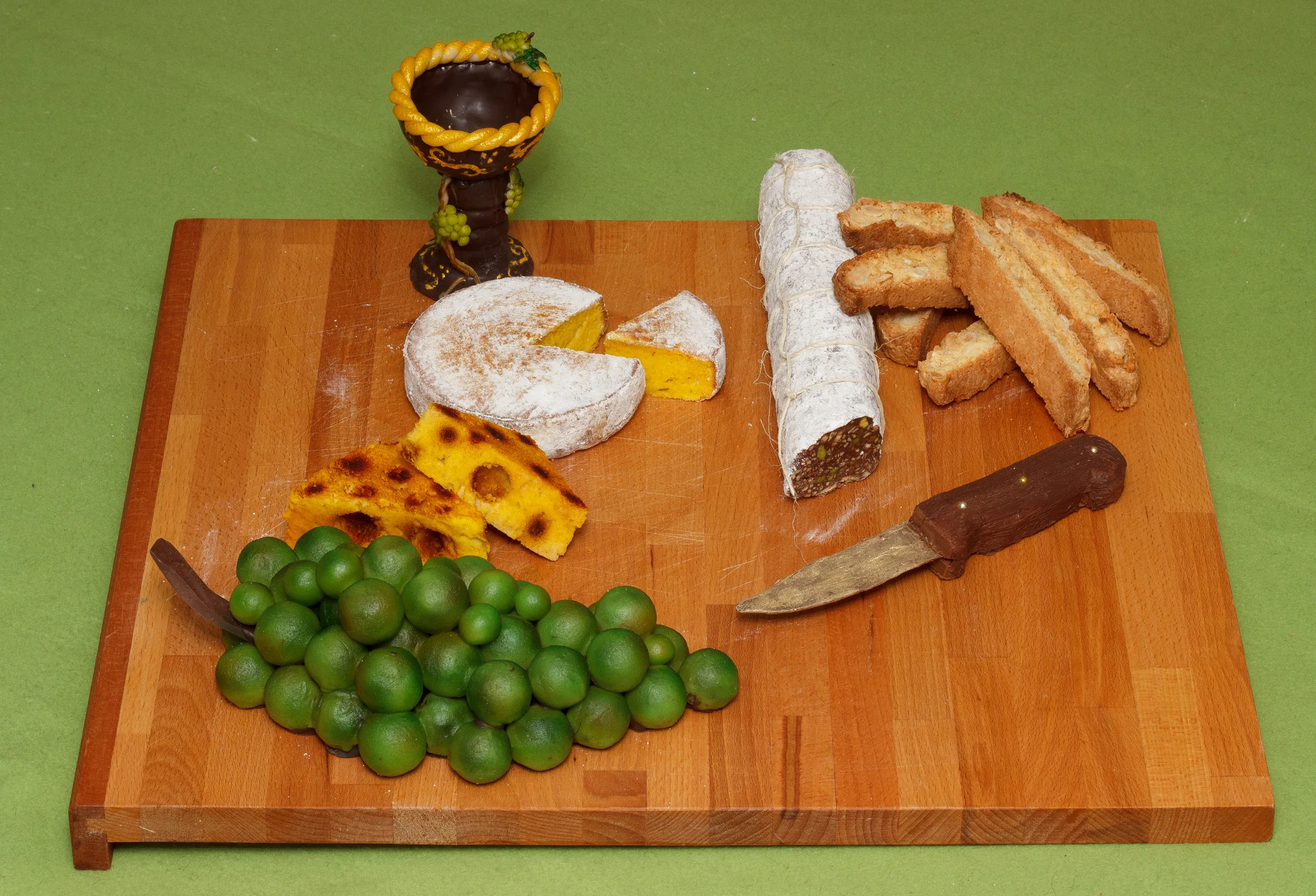 Holzbrett mit einer Vesperplatte, bei der alles aus Süßem besteht: Käselaib, Salami, Weintrauben, Cantuccini und ein Messer aus Schokolade
