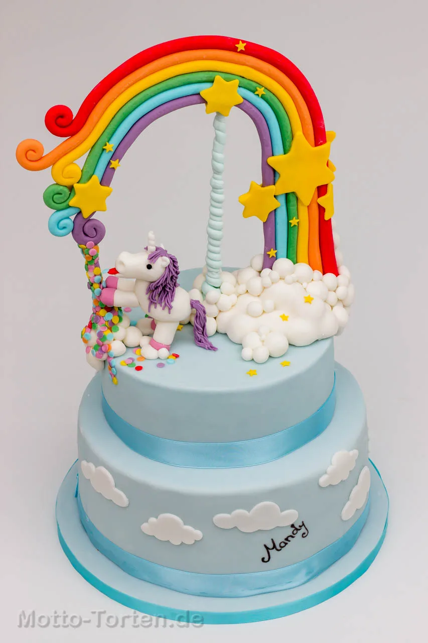 Zweistöckige hellblaue Geburtstagstorte mit Wolken aus Fondant, darauf ein modelliertes Einhorn unter einem großen Regenbogen mit Sternen und Konfetti