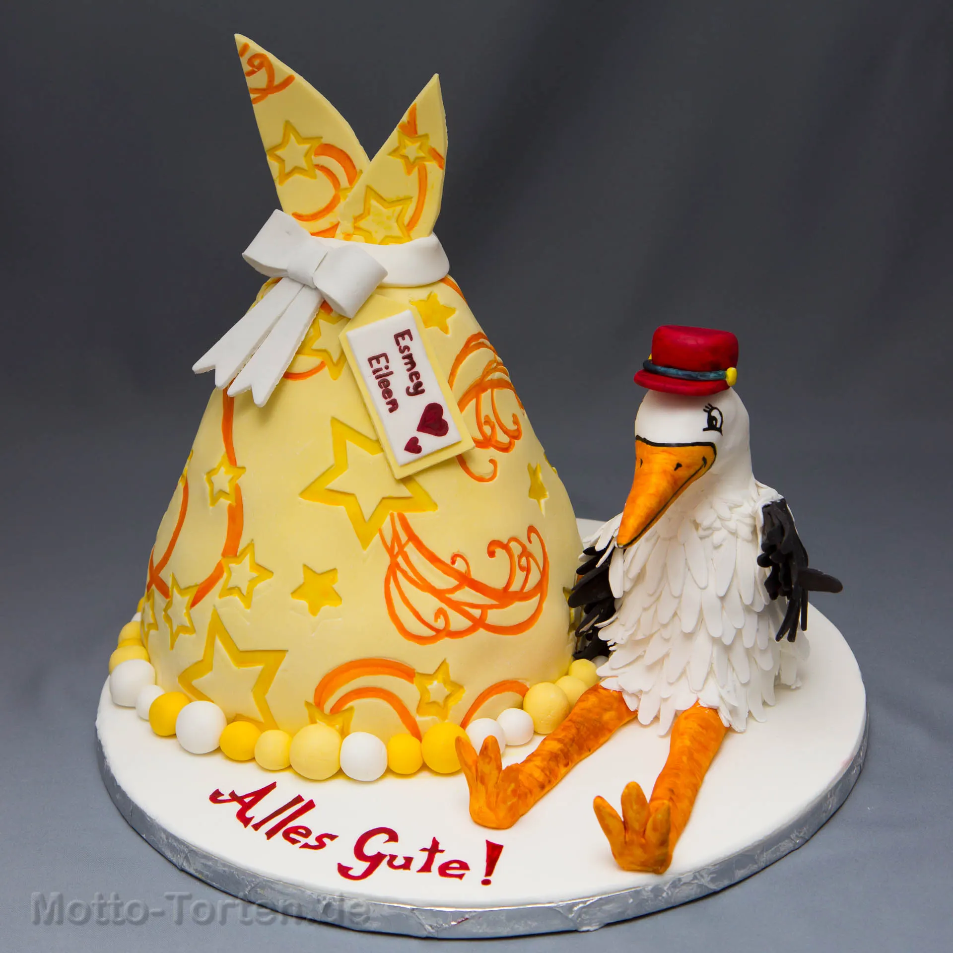 Torte in Form eines gelben Storchenbündels mit Sternen, Schleife und Namensschild, daneben ein Klapperstorch aus Fondant mit rotem Hut