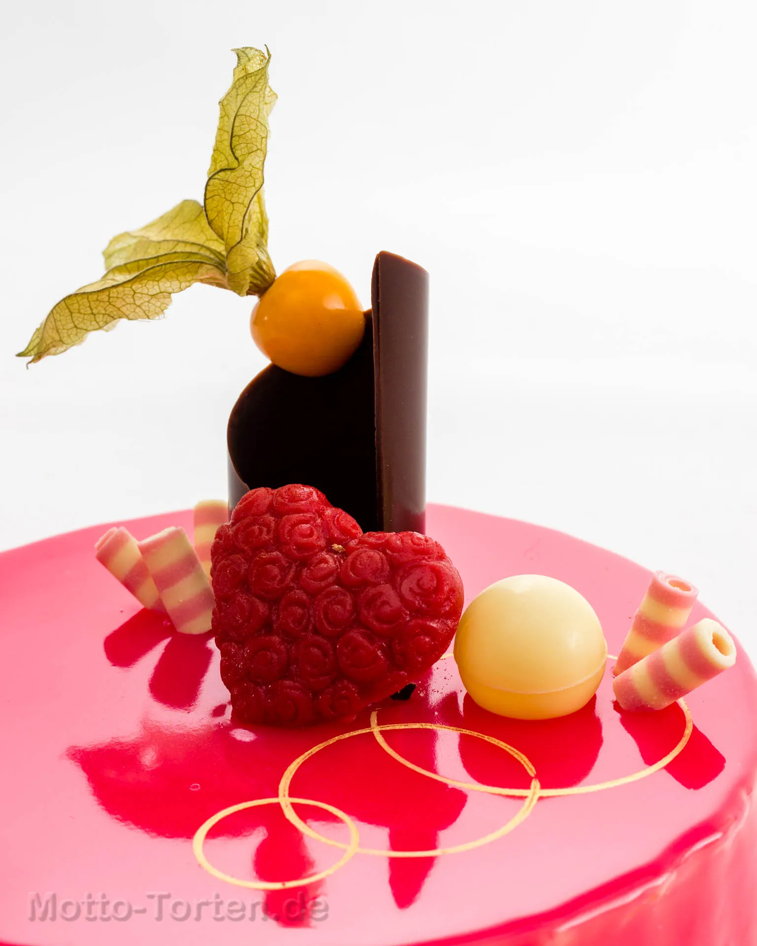 Torte mit pinkfarbener Spiegelglasur, darauf ein Herz aus Himbeermasse, eine weiße Schokoladenkugel, Physalis und gestreifte Schokoröllchen