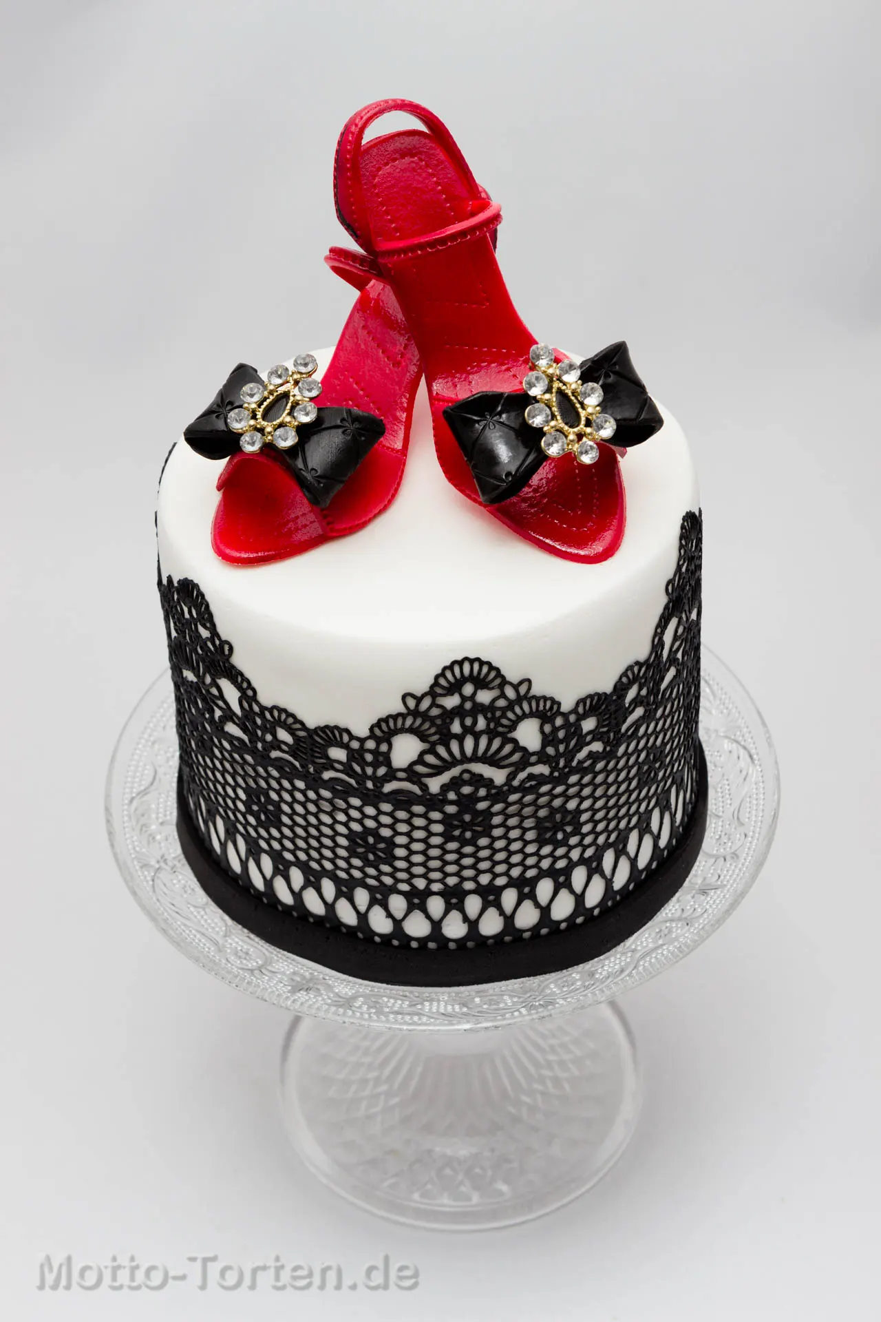 Weiße Fondanttorte mit schwarzer essbarer Spitze, obenauf zwei rote High Heels aus Fondant mit schwarzen Schleifen und Strasssteinen