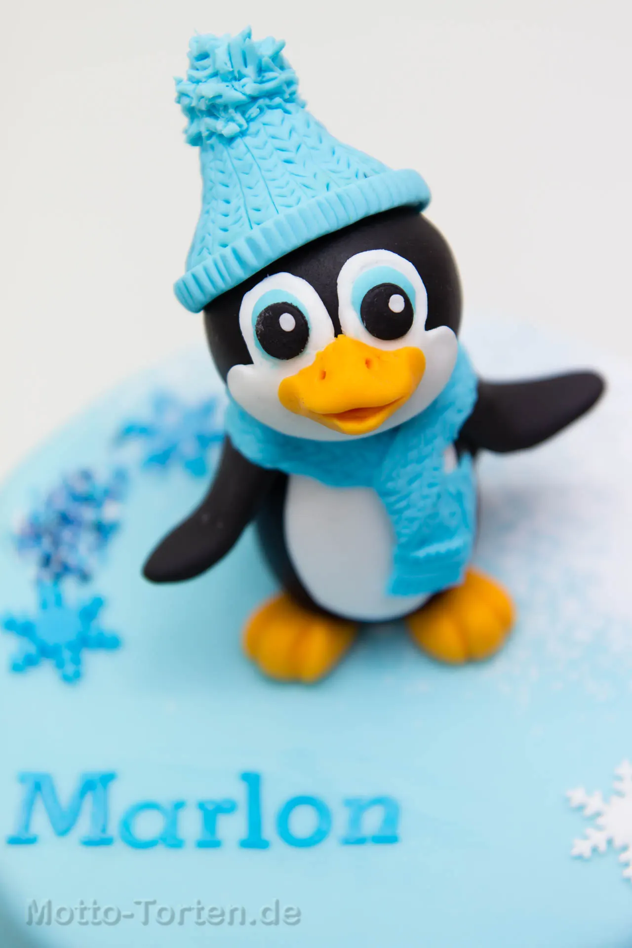 Handmodellierter Pinguin mit blauer Strickmütze und Schal auf einer hellblauen Tauftorte, daneben Schneeflocken und der Name Marlon