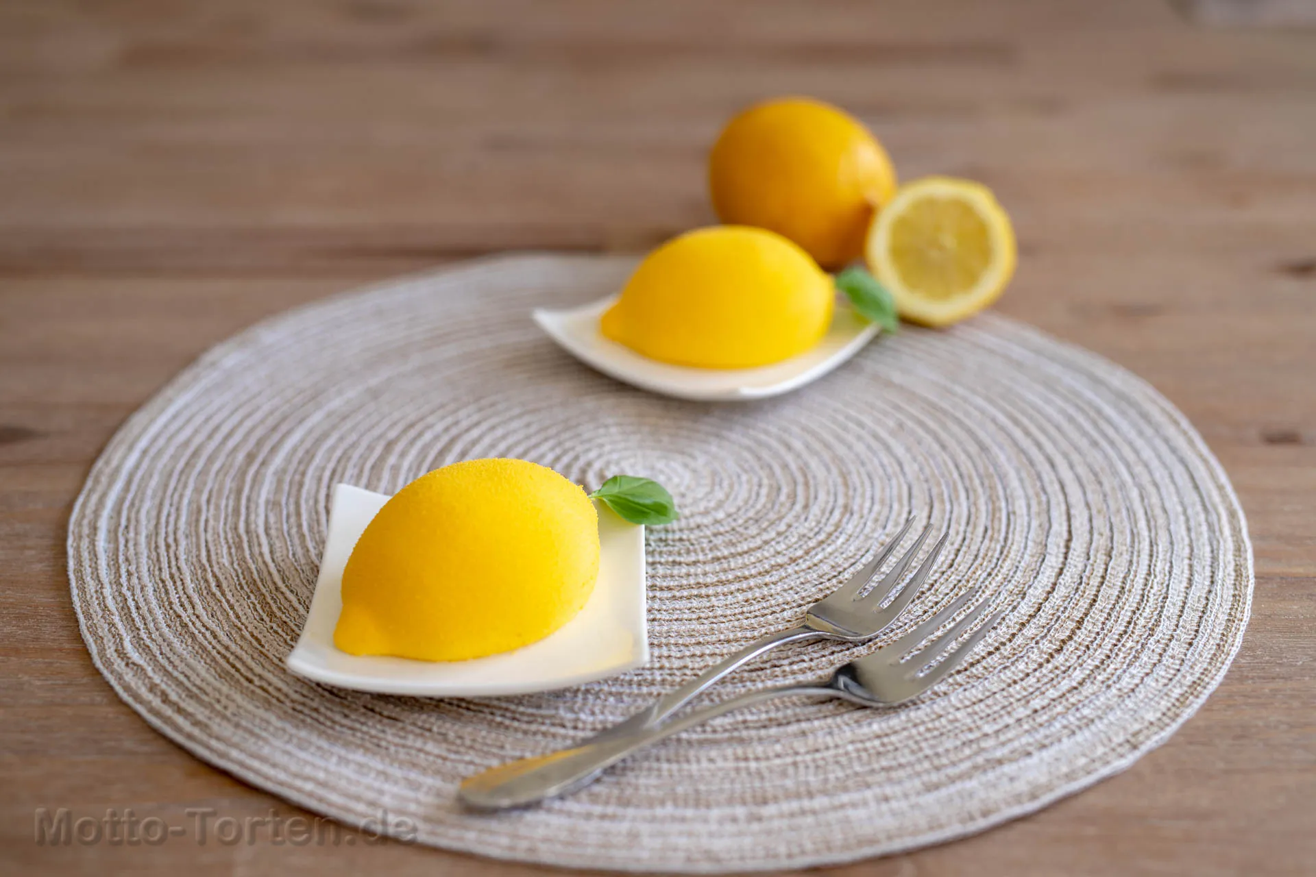 Zwei zitronenförmige Törtchen mit samtig gelber Oberfläche auf weißen Schälchen, daneben frische Zitronen und Kuchengabeln