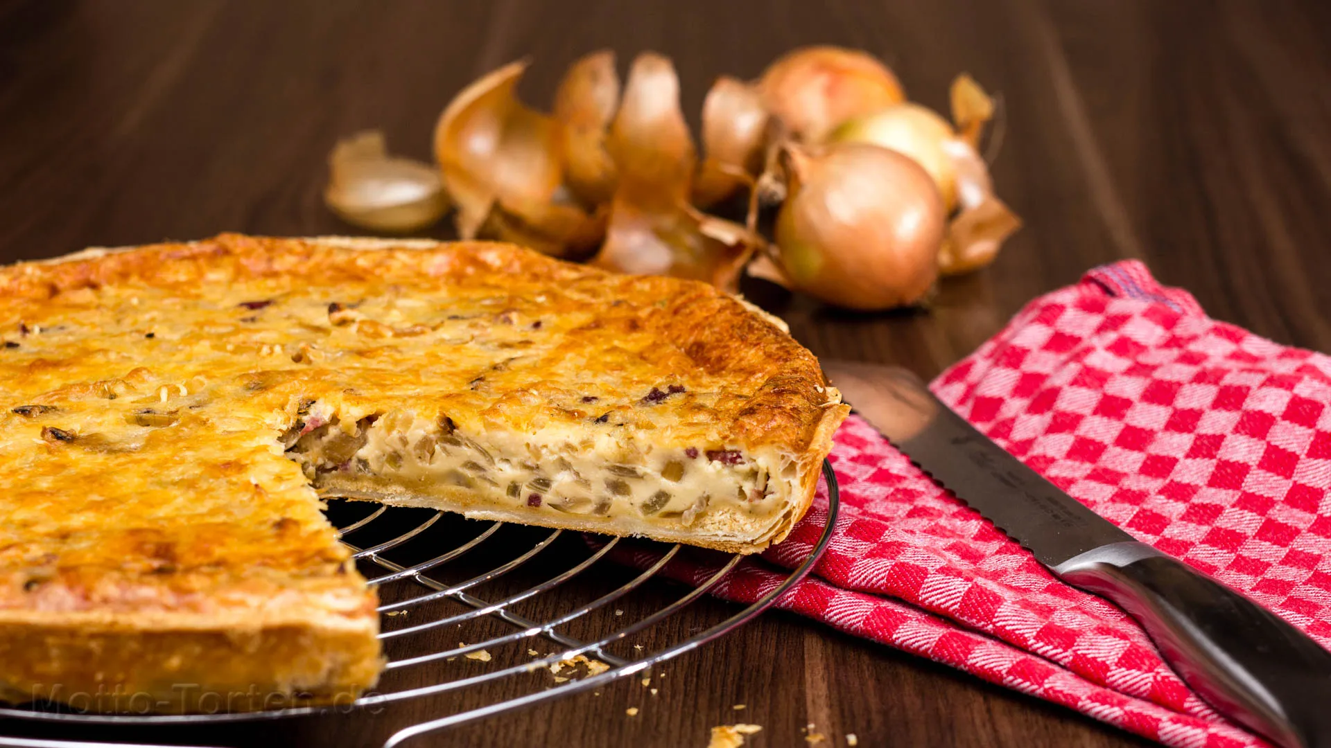 Angeschnittener Zwiebelkuchen auf einem Kuchengitter, daneben Zwiebeln und ein Messer auf rot kariertem Tuch