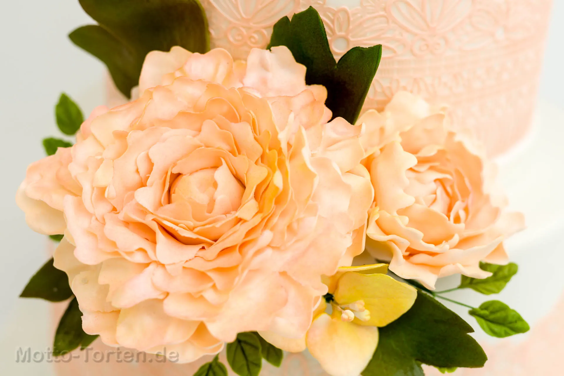 Mehrstöckige weiße Hochzeitstorte mit apricotfarbener essbarer Spitze und handmodellierten Pfingstrosen in Apricot mit grünen Blättern – Ansicht 2