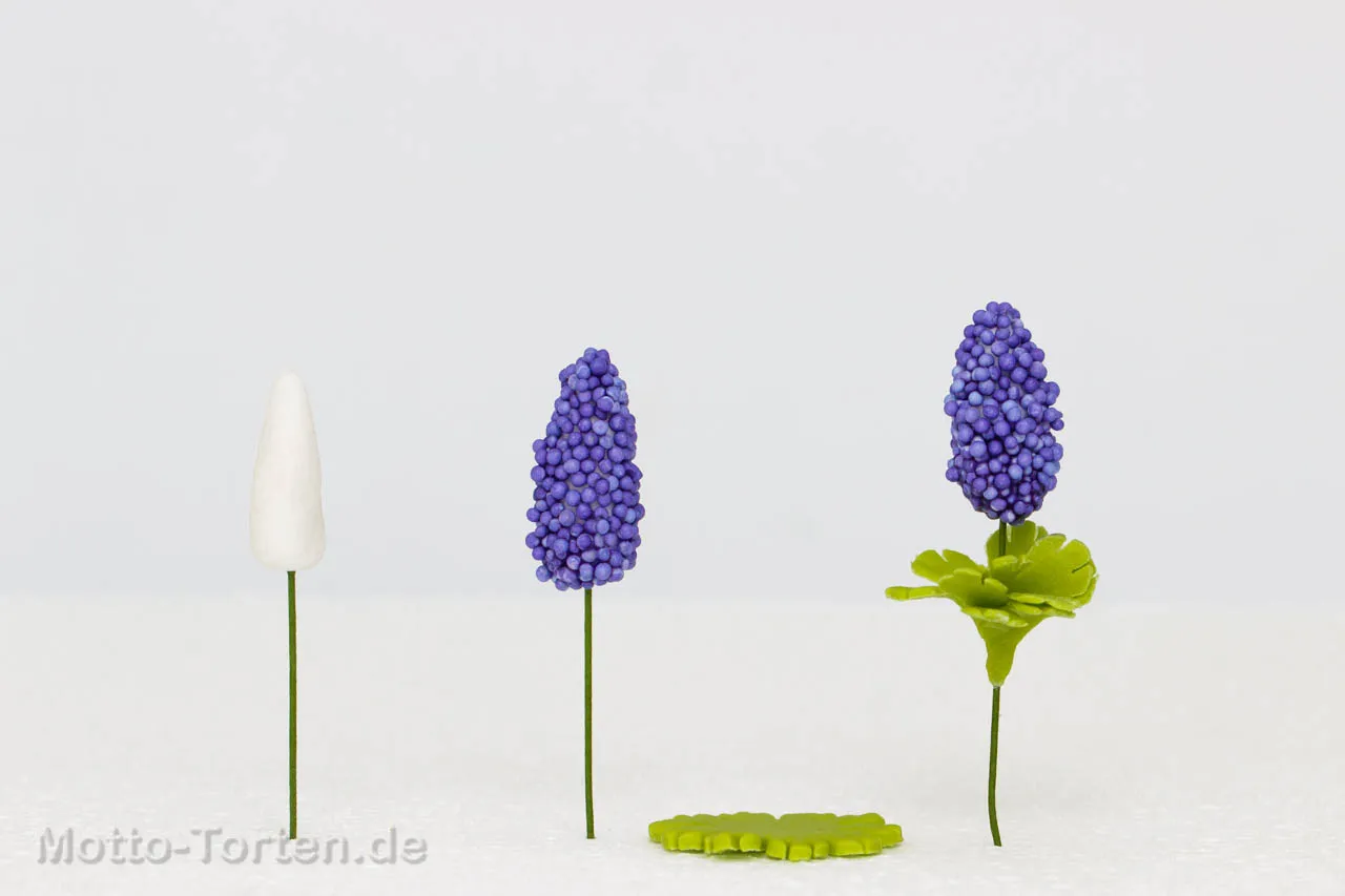 Drei Arbeitsstände nebeneinander: weißer Tropfen am Draht, in blauen Nonpareilles gerollt, fertige Blüte mit grünen Blättern