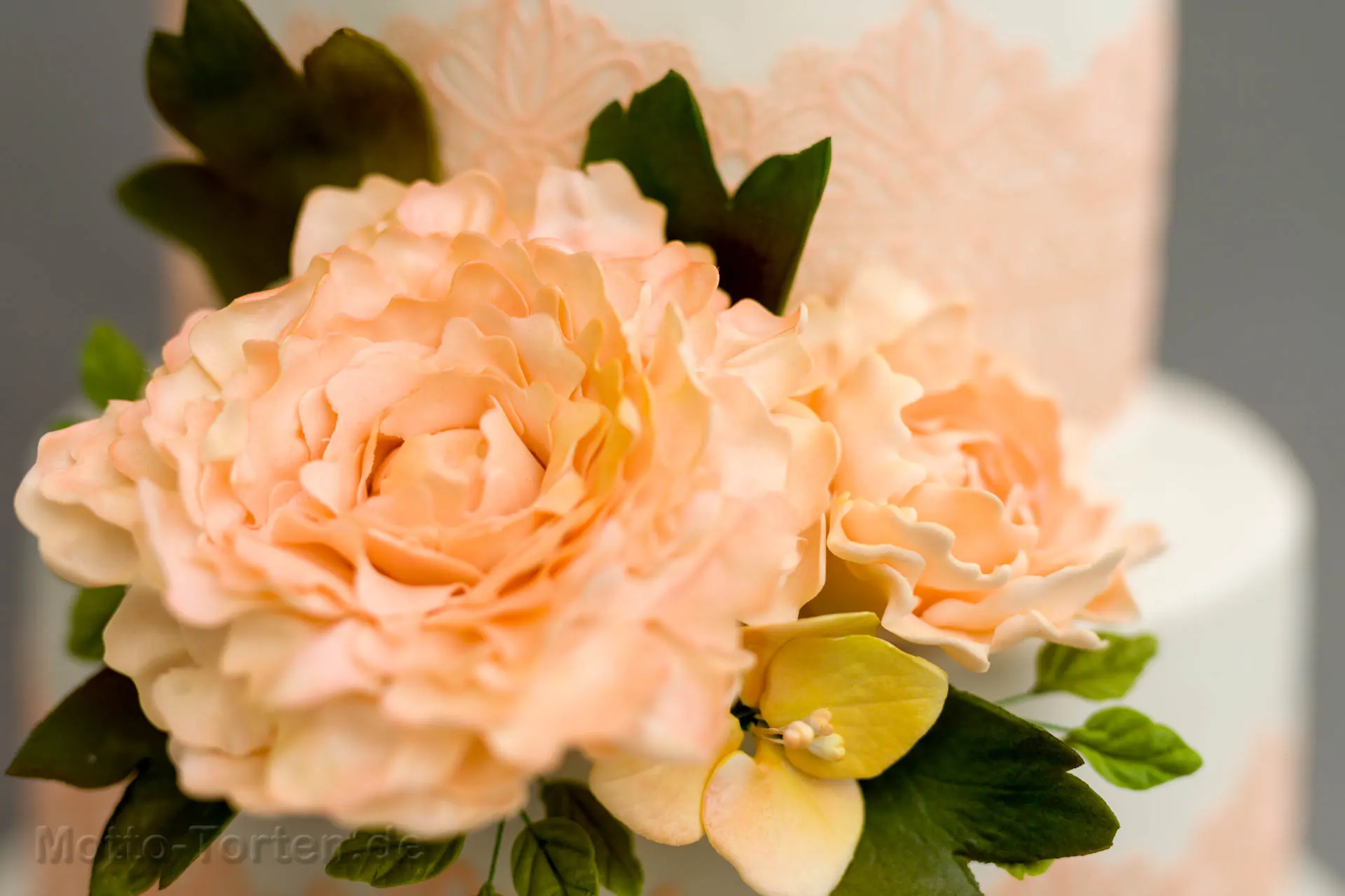 Mehrstöckige weiße Hochzeitstorte mit apricotfarbener essbarer Spitze und handmodellierten Pfingstrosen in Apricot mit grünen Blättern – Ansicht 4