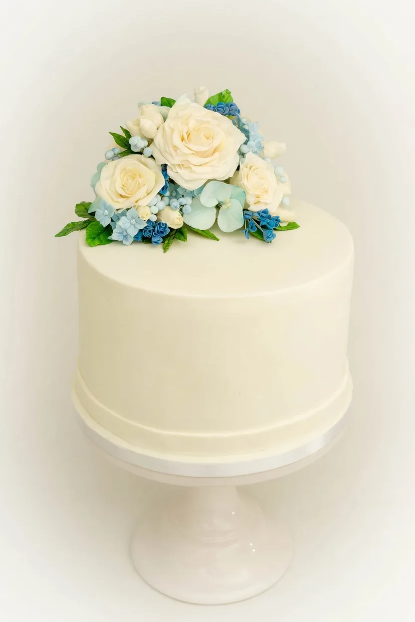 Elegante einstöckige Hochzeitstorte in Creme mit handgefertigten weißen und blauen Zuckerrosen auf einer Tortenetagere, fotografiert im Tortenatelier
